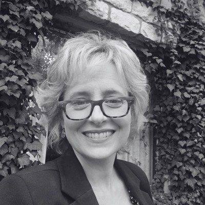 Smiling person with glasses outdoors in front of ivy-covered wall.