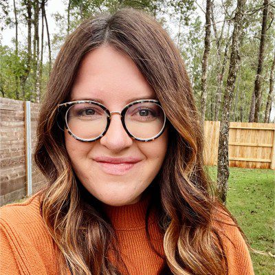 A smiling woman with glasses and wavy hair outdoors.